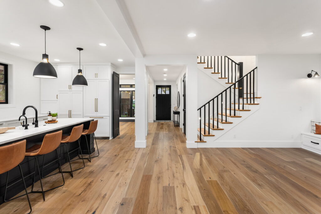 Kitchen and living room interior in new luxury home with open concept floor plan. Features large island with farmhouse sink, hardwood floors, and stairs to second story.